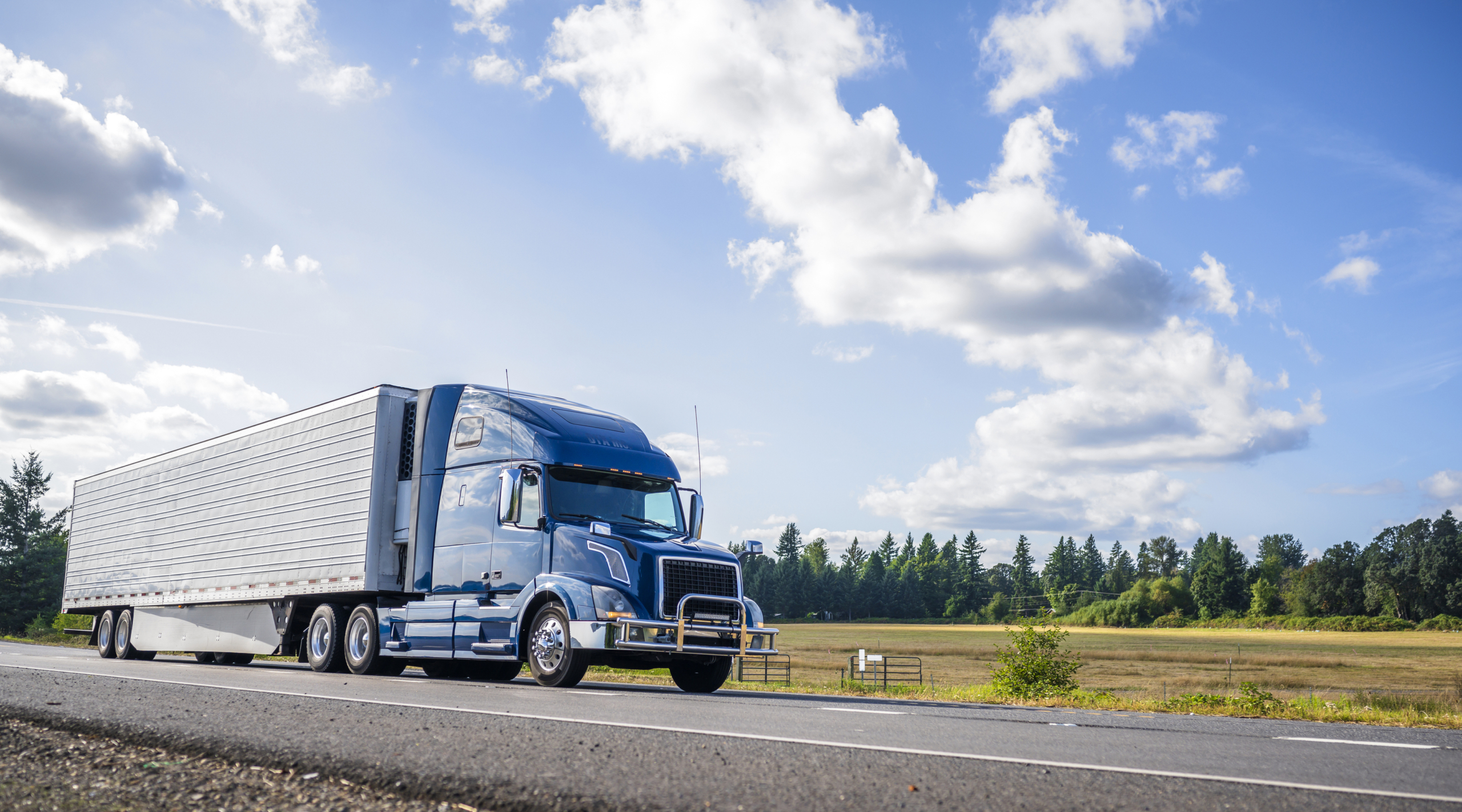 Big rig powerful professional industrial blue bonnet semi truck for long haul delivery commercial cargo going with refrigerator semi trailer on the summer road with forest and meadows on the sides