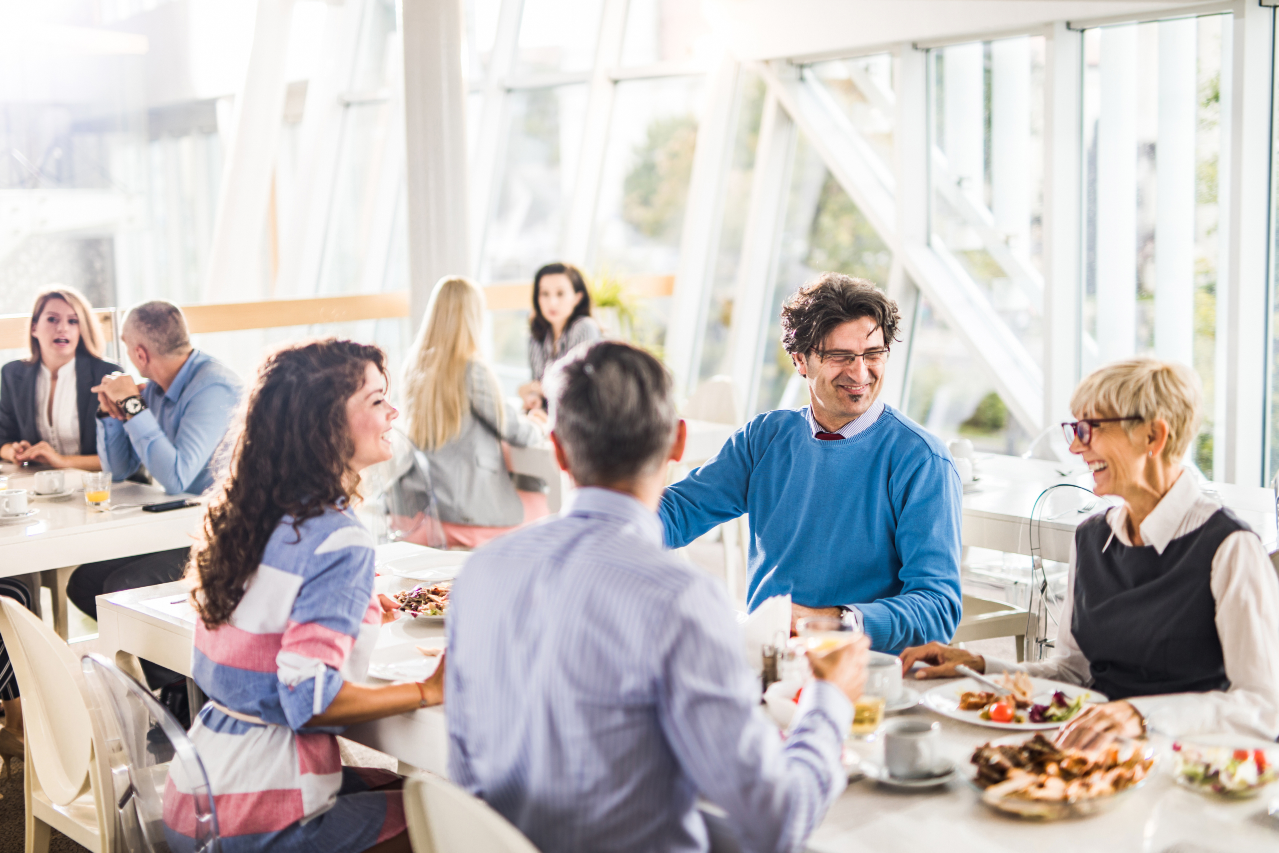 Group of happy business people communicating while having lunch break in cafeteria. Focus is on man in blue sweater.