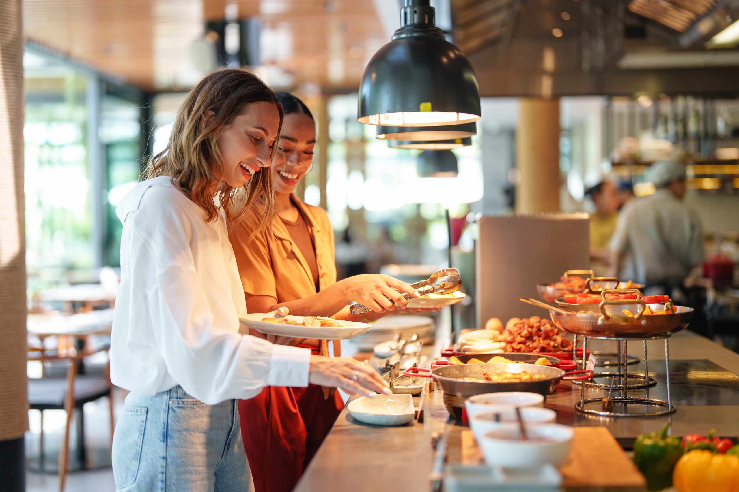 Young adult multiracial and Caucasian females smile while selecting food in a self-service restaurant. Both dressed casually, they enjoy a variety of dishes under warm lighting, creating a friendly and relaxed ambiance.